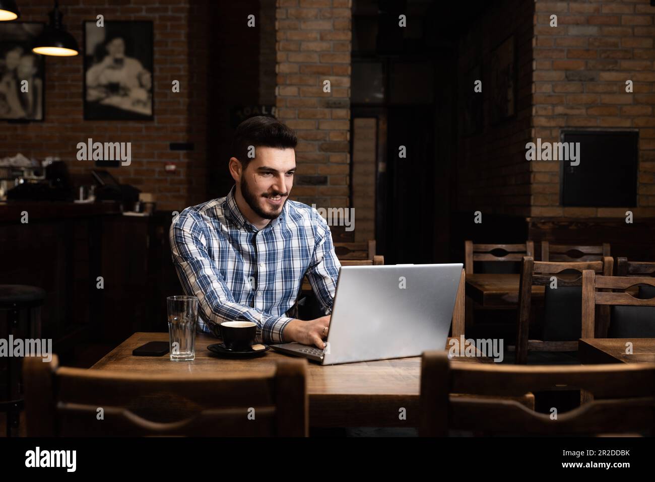 Ein junger Geschäftsmann, der Inhaber oder Manager einer gemütlichen Bar und Cafeteria ist, sitzt nach Ende der Arbeitszeit und kontrolliert die Einnahmen und den Umsatz über ein Notebook Stockfoto