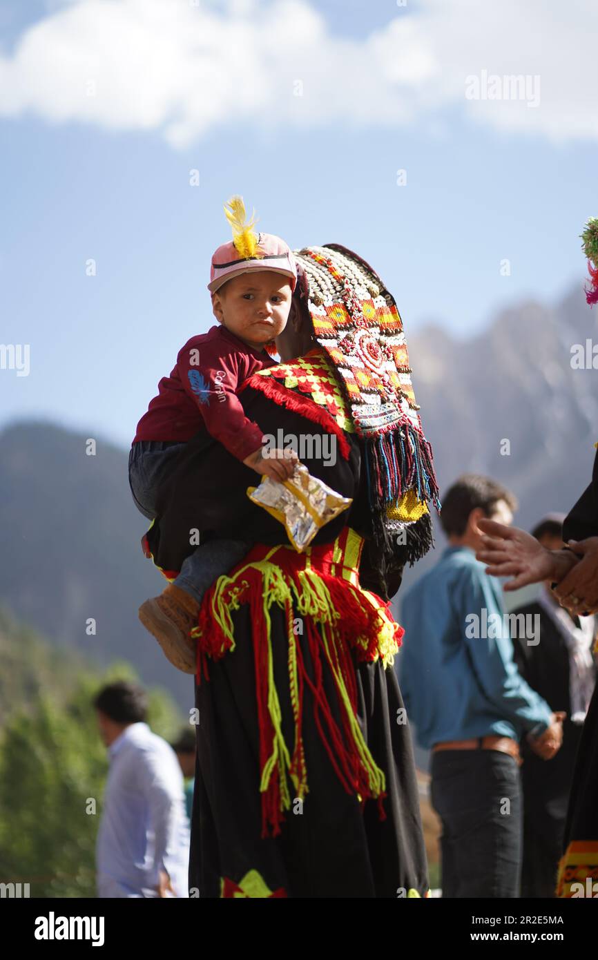 Bamburet, KPK, Pakistan - 05152023: Eine Kalash-Frau kleidet sich in traditioneller Kleidung und hält ein Kind, während sie das Chilam Joshi Festival in Chitral betrachtet Stockfoto