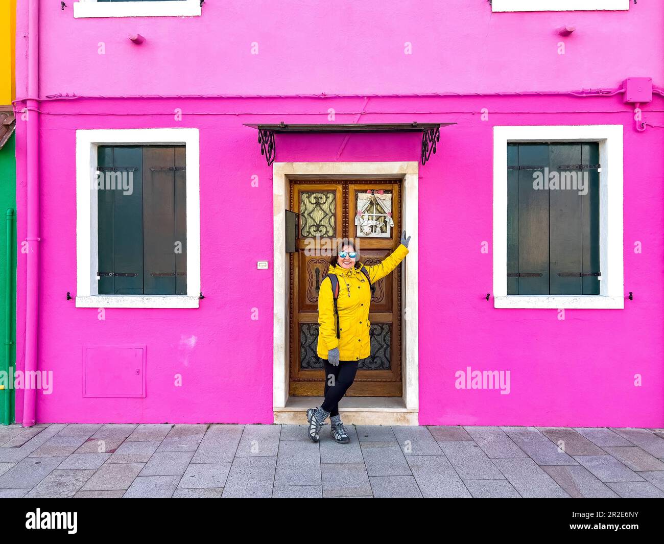 Lächelnde Frau, die vor dem rosa Haus auf Burano Island posiert. Glückliche Reisende, weibliche Touristen mit gelbem Regenmantel und Sonnenbrille, posieren und Spaß haben Stockfoto