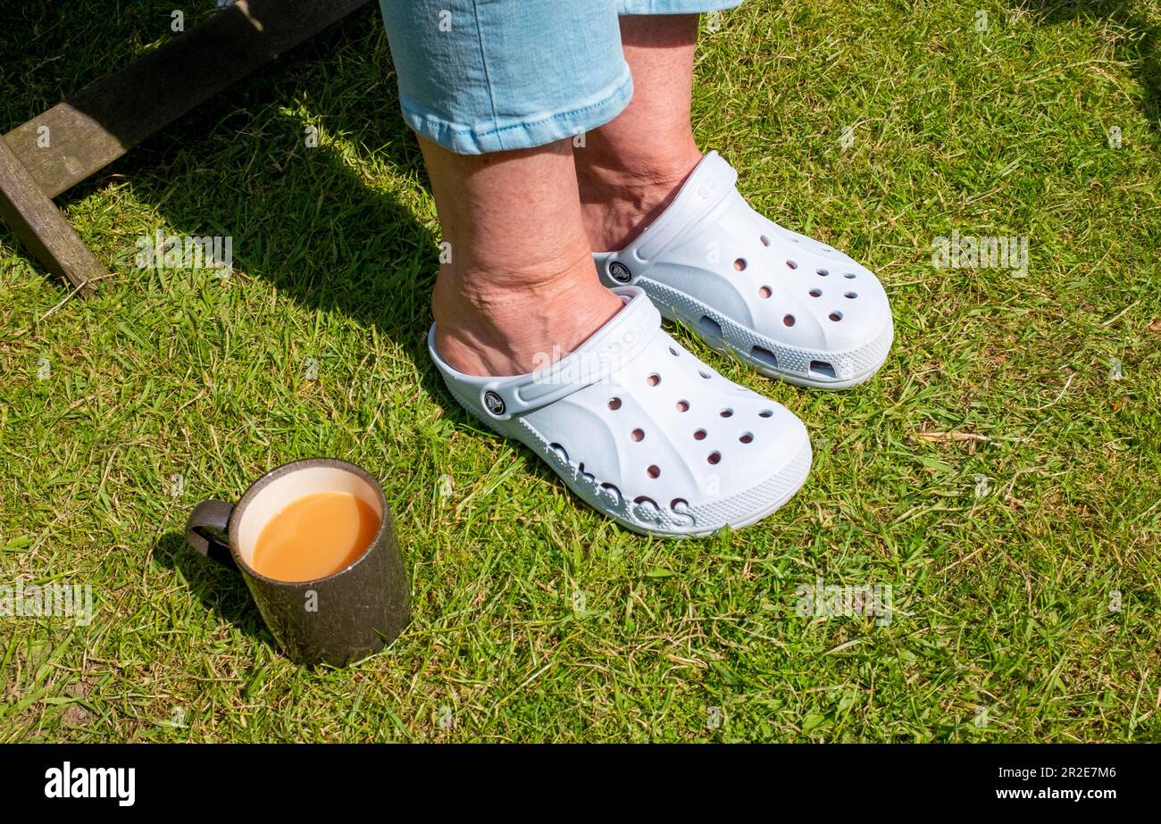 Frau trägt ein Paar Crocs-Schuhe zu einer Tasse Tee in einem städtischen Garten während der Frühlingssonne Stockfoto