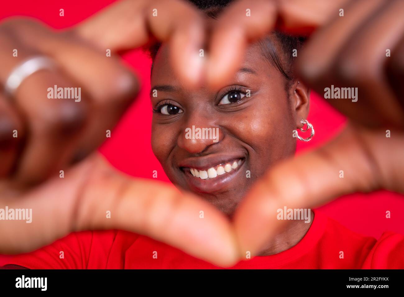 Junge afroamerikanische Frau, isoliert auf rotem Hintergrund, lächelnd und Herzgesten, Studioaufnahme Stockfoto