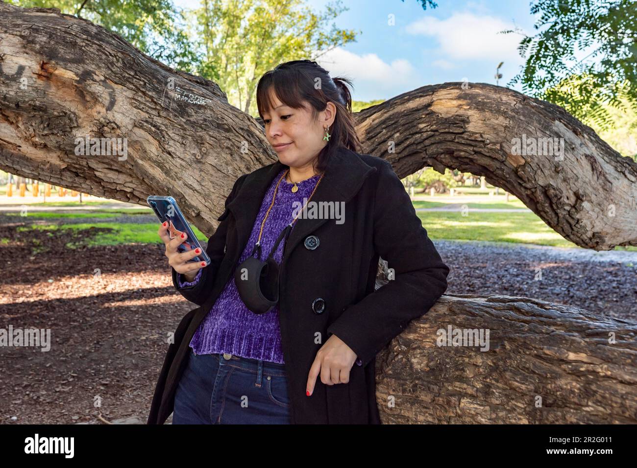 Eine Frau liest eine SMS auf ihrem Handy, während sie sich an einen Baum im Park lehnt Stockfoto