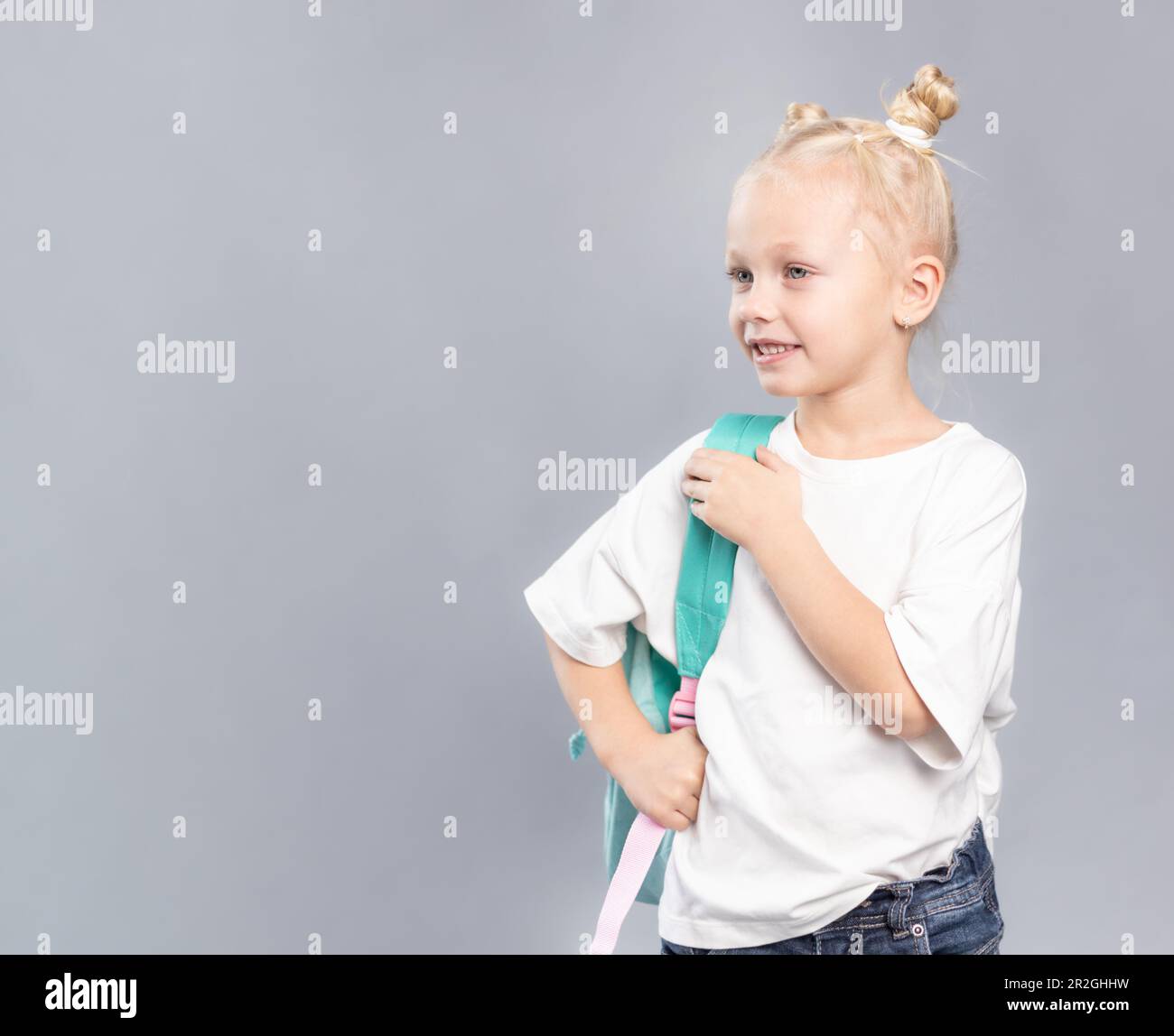 Ein Mädchen der Grundschule lächelt und hält einen Rucksack, ein Kind mit blonden Haaren und dreckigen Brötchen, gekleidet in einem weißen T-Shirt, das seitlich auf grauem Hintergrund aussieht Stockfoto