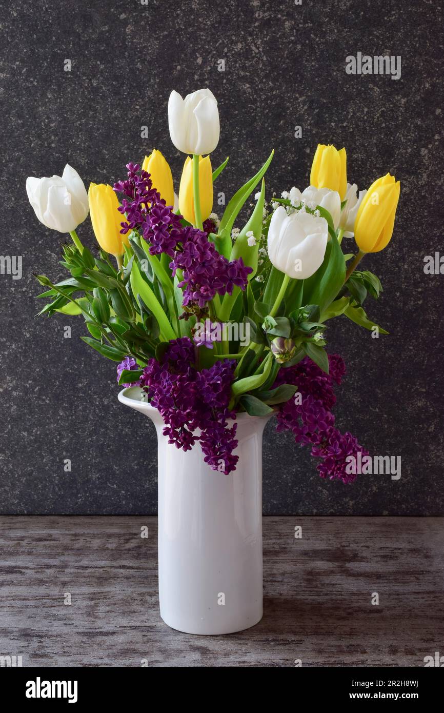 Ein wunderschöner Strauß aus Tulpen und Fliedern in einer weißen Vase vor dunklem Steinhintergrund, Seitenansicht Stockfoto