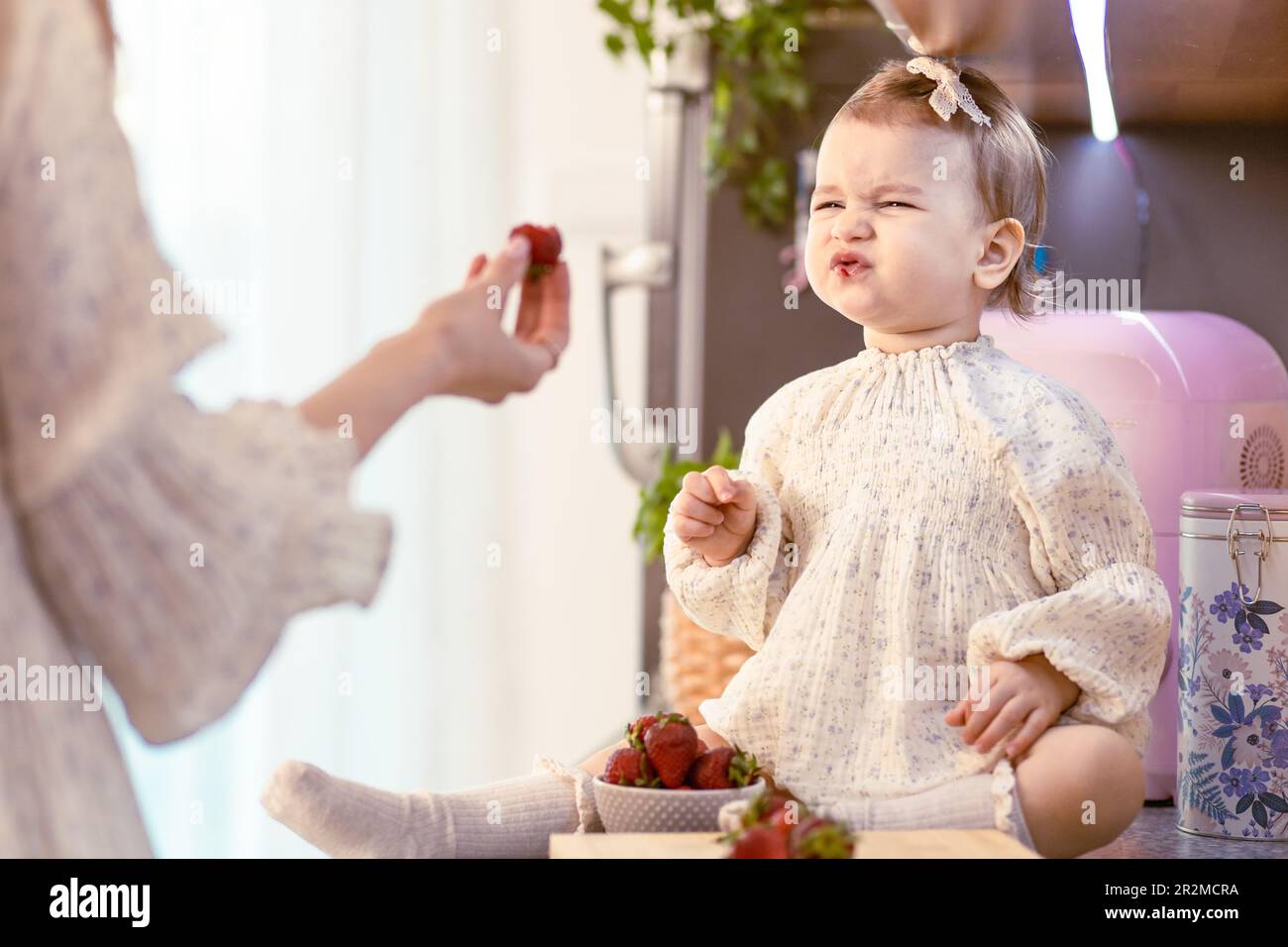 Mom füttert ihr kleines Mädchen mit sauren Erdbeeren in der Küche. Du machst ein sauriges Gesicht Stockfoto