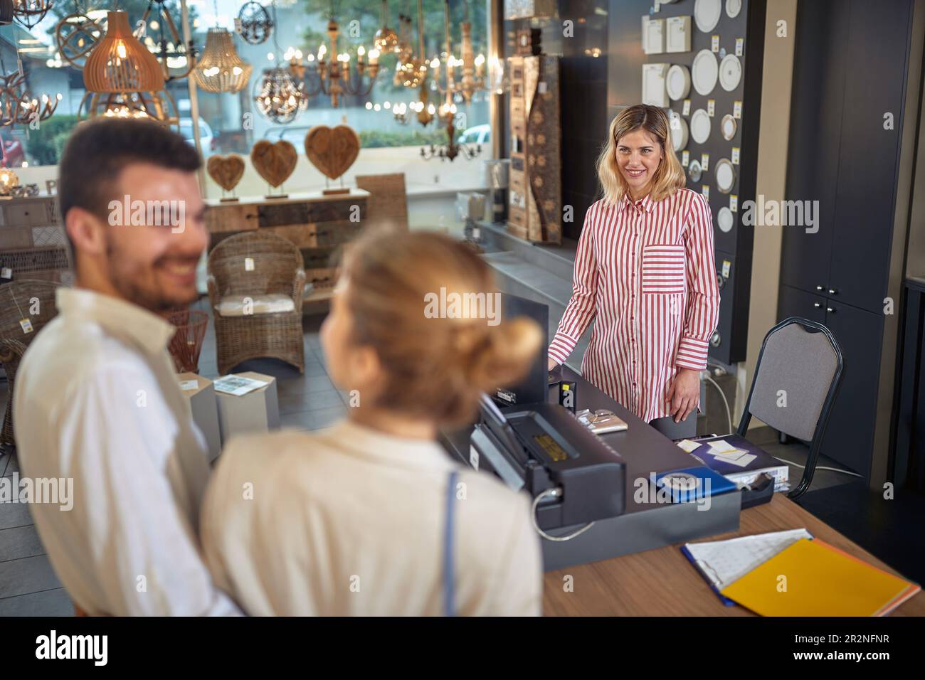 Eine reizende junge Verkäuferin, die ein junges Ehepaar im Lampengeschäft bedient. Ein Paar kauft Inneneinrichtung und Lampen. Einkaufen, Lifestyle, Zusammensein Conc Stockfoto