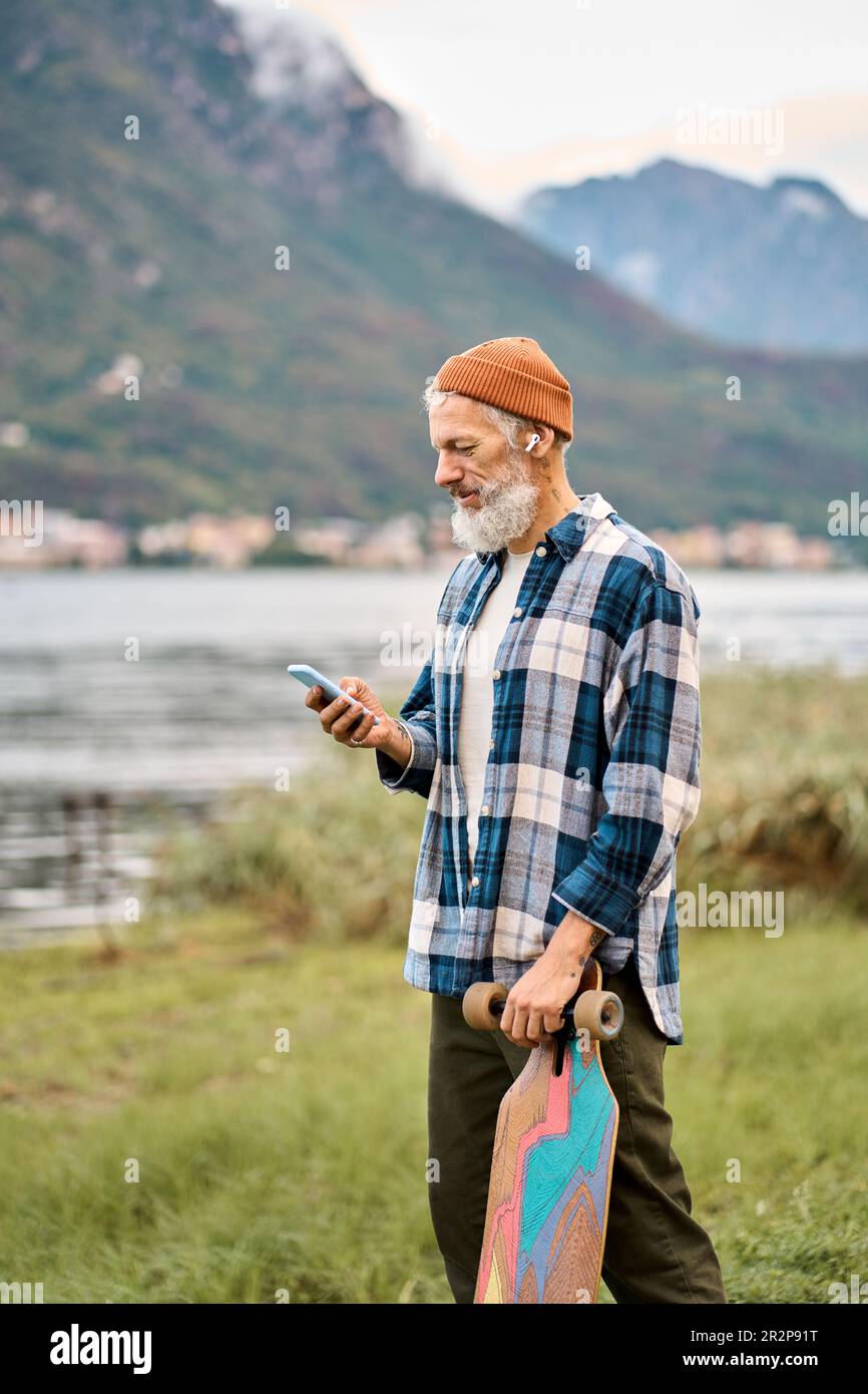 Aktiver alter Hipstermann, der im Naturpark steht und Skateboard am Telefon hält. Stockfoto