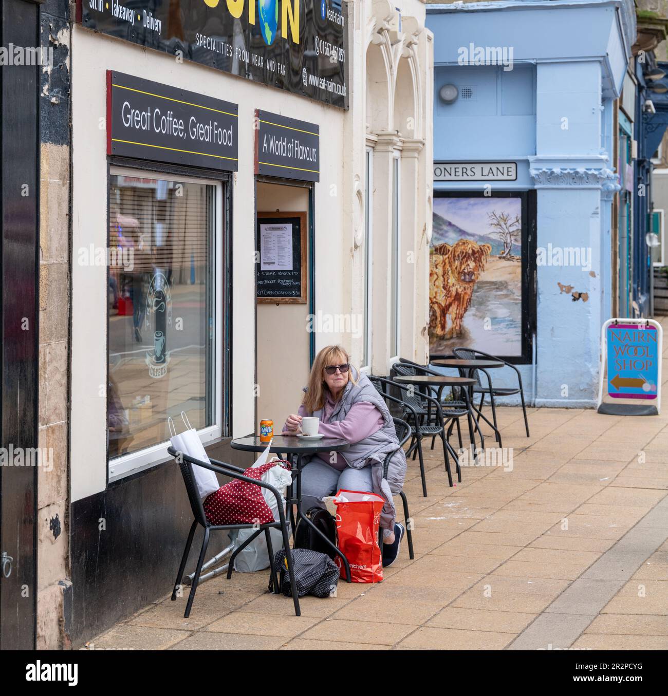 20. Mai 2023 High Street, Nairn, Schottland. Das ist eine Frau, die vor einem Café eine Tasse Kaffee trinkt und von einer Highland Cow beobachtet wird Stockfoto