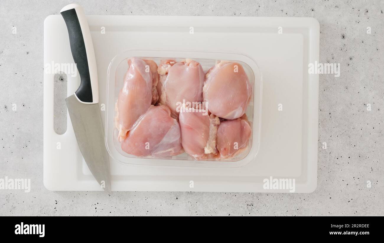 Hähnchenschenkel in Kunststoffbehälter und Küchenmesser Nahaufnahme auf einem weißen Kunststoffschneidbrett auf einem Küchentisch. Mariniertes Hühnerfilet-Rezept, Stockfoto