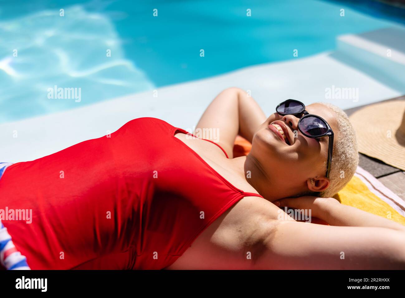Fröhliche, modische, unveränderte, birassische Frau in Sonnenbrille und Badeanzug, Sonnenbaden am Pool, lächelnd. Sommer, Freizeit, Entspannung und Urlaub. Stockfoto