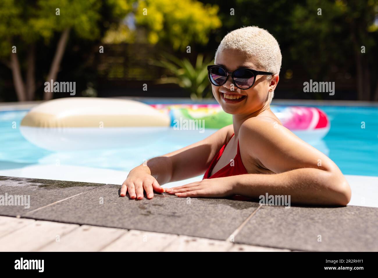 Porträt einer glücklichen, unveränderten, birassischen Frau mit Sonnenbrille, die am Pool steht. Sommer, Freizeit, Entspannung und Urlaub. Stockfoto