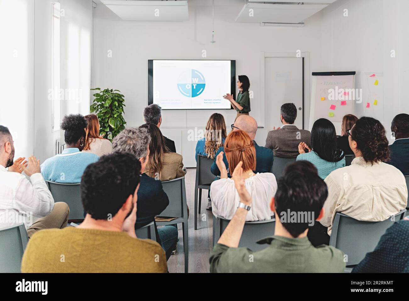 Geschäftsfrau präsentiert SWOT-Analyse auf einem großen Bildschirm in einem geräumigen Büroraum. Vielfältige Zuschauer von hinten gesehen, klatschend in die Hände. Firmeneinrichtung. Stockfoto