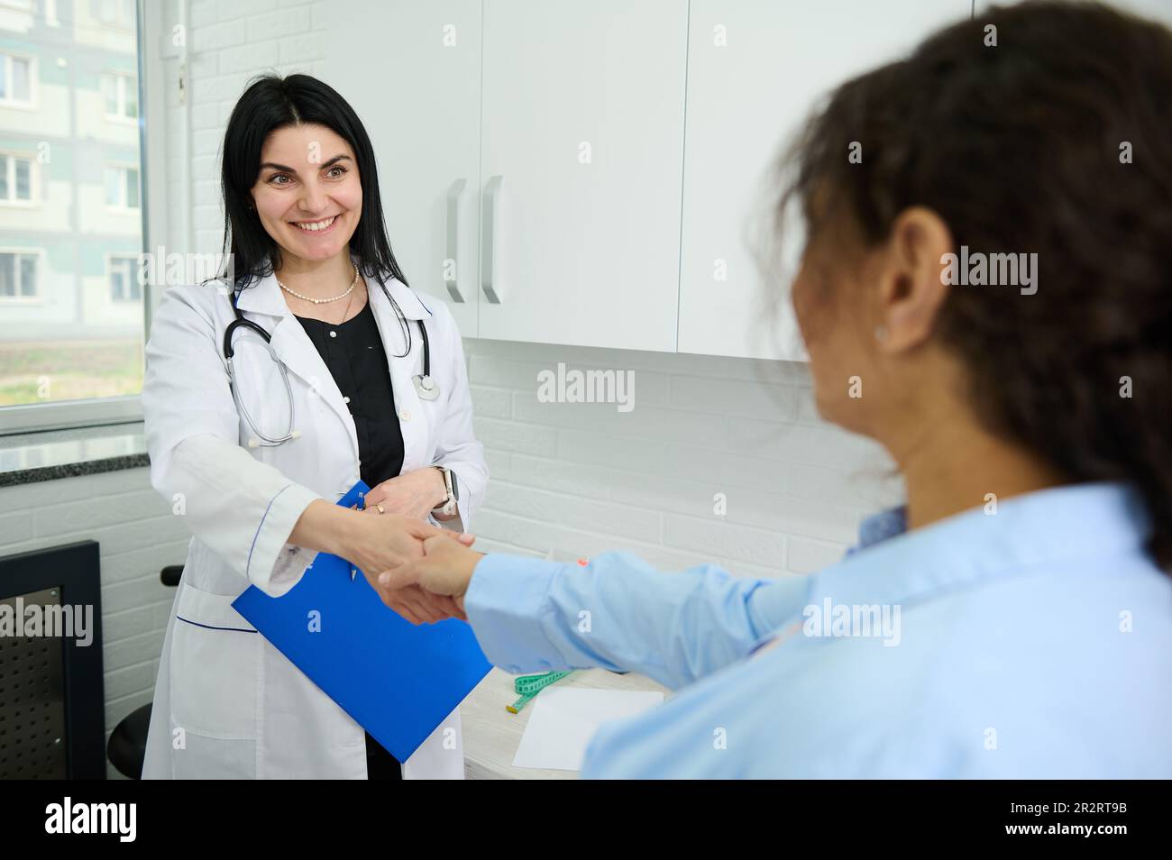 Eine selbstbewusste, weiße Ärztin, die ein Klemmbrett mit einem medialen Versicherungsvertrag hält, lächelt, schüttelt dem neuen Patienten die Hand Stockfoto Eine selbstbewusste, weiße Ärztin, die ein Klemmbrett mit einem medialen Versicherungsvertrag hält, lächelt, schüttelt dem neuen Patienten die Hand Stockfoto