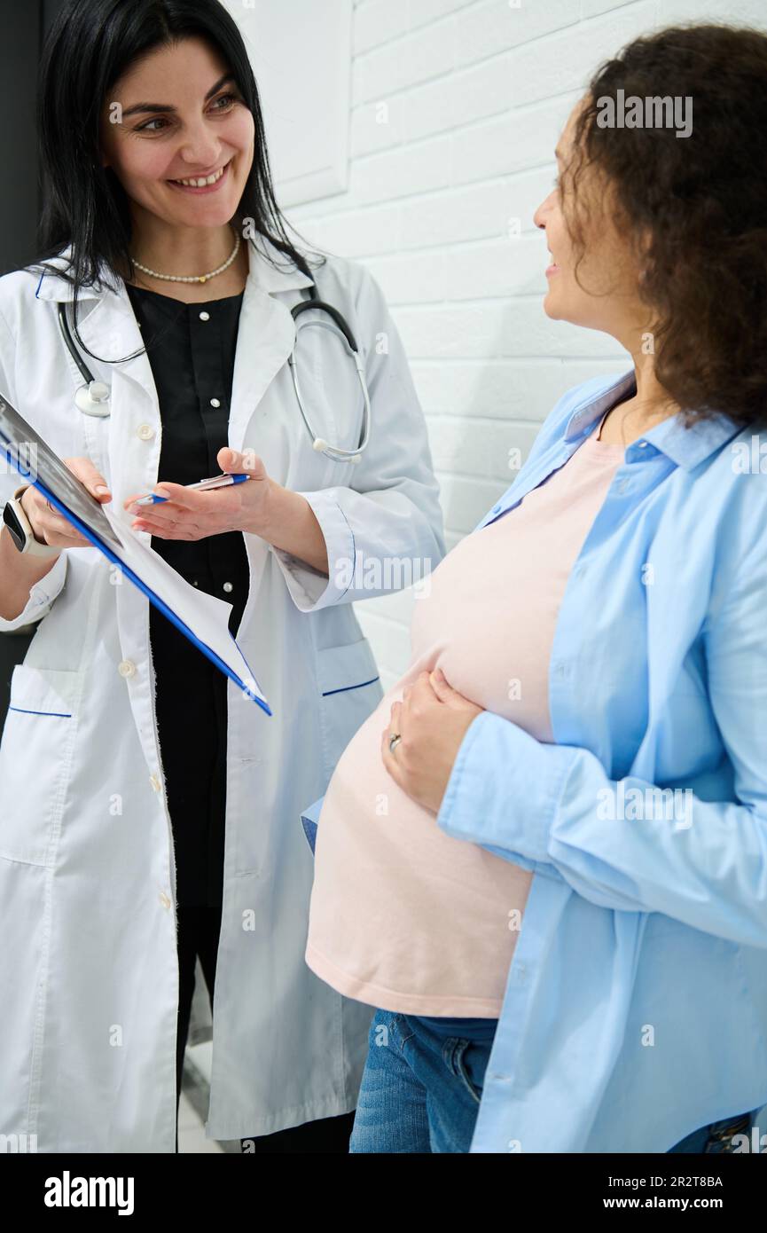 Weibliche Gynäkologin hält ein Klemmbrett, erklärt schwangeren Frauen die Behandlung, die in der Geburtshilfe-Klinik durchgeführt werden muss Stockfoto
