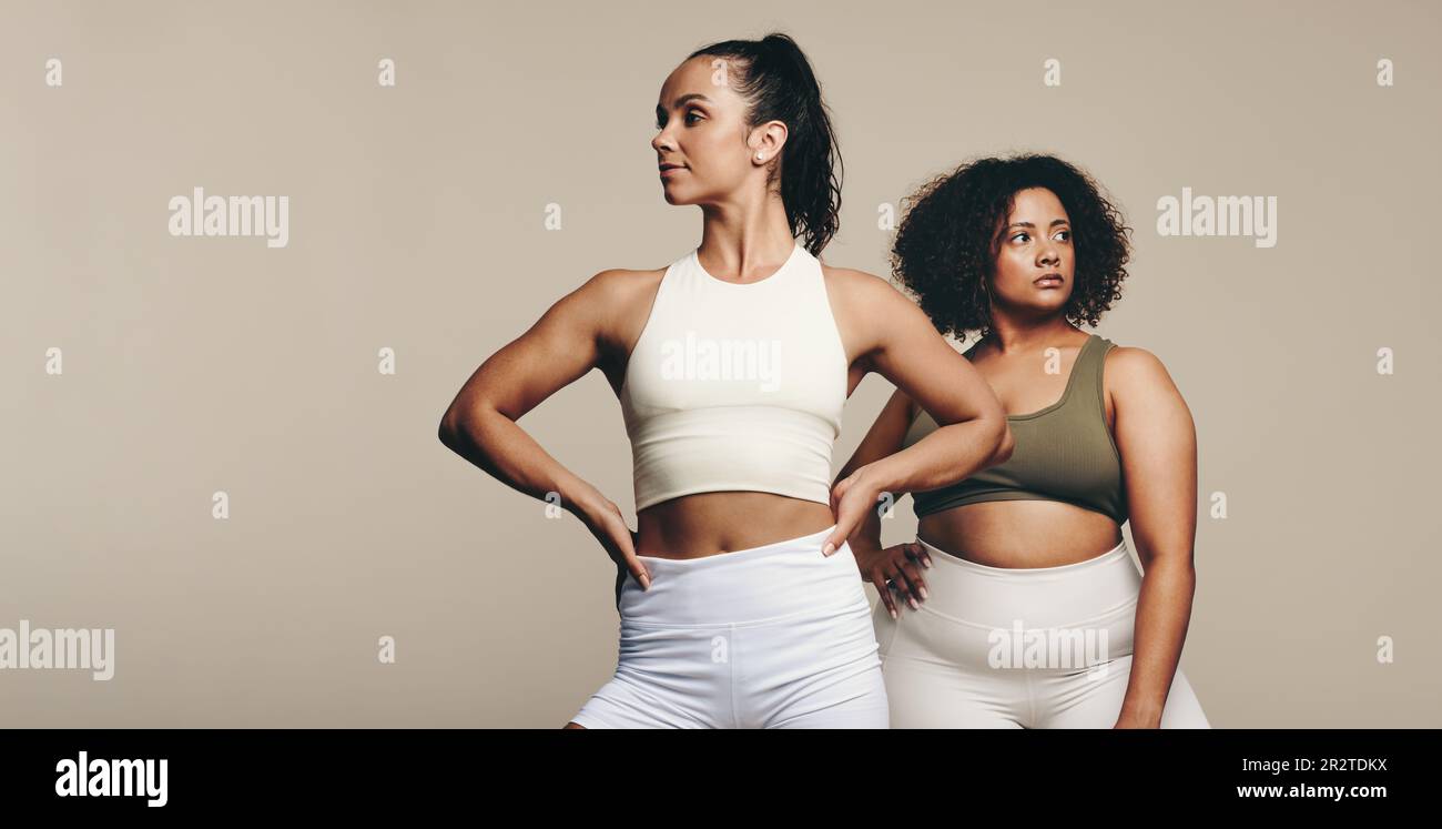 Passen Sie Frauen mit unterschiedlichen Körperformen an, die in Sportbekleidung zusammen in einem Studio stehen. Zwei Sportlerinnen zeigen ihren Körper, während sie umarmen Stockfoto