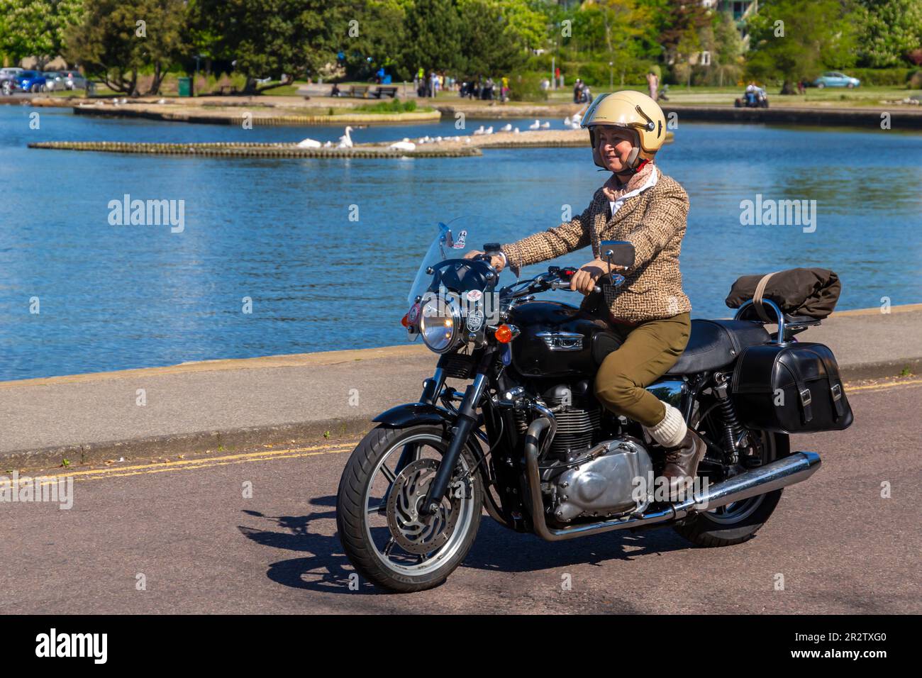 Poole, Dorset, Großbritannien. 21. Mai 2023 Bournemouth and Poole Distinguished Gentleman’s Ride ist eine von vielen weltweit jährlich stattfindende Veranstaltung, um Geld und Bewusstsein für die Gesundheit von Männern zu schaffen. Im Rahmen der Fahrt fahren Hunderte von Motorrädern, viele davon im Vintage-Stil, durch den Poole Park auf dem Weg nach Bournemouth mit vielen ihrer Fahrer, die angemessen gekleidet sind, in Krawatten im Retro-Stil, Schnurrbart und Tweeds an einem schönen sonnigen Tag. Frau, die Jacke und Schal trägt und Triumph Motorrad fährt. Quelle: Carolyn Jenkins/Alamy Live News Stockfoto