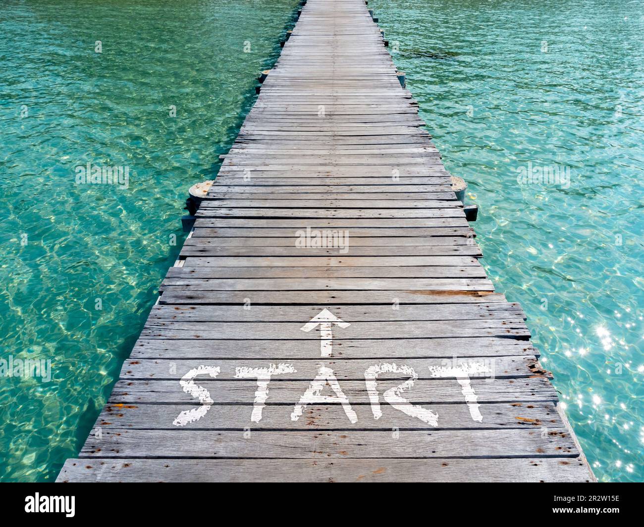 Start, weißer Text und Pfeil auf der Holzbrücke Richtung Meer. Holzplank Pathway Bridge auf klarem Meerwasser am Hafen der Insel an sonnigen Tagen. Begi Stockfoto