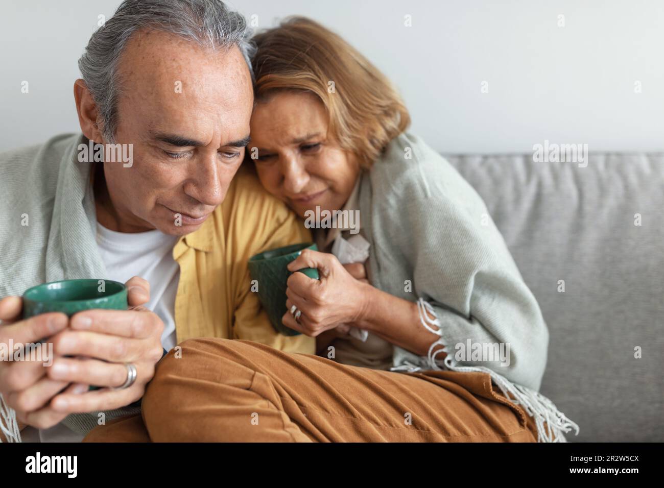 Altes Paar, das in eine Decke eingewickelt sitzt und Tassen mit heißem Tee oder Kaffee hält, sich krank und kalt fühlt, zu Hause auf dem Sofa sitzt Stockfoto