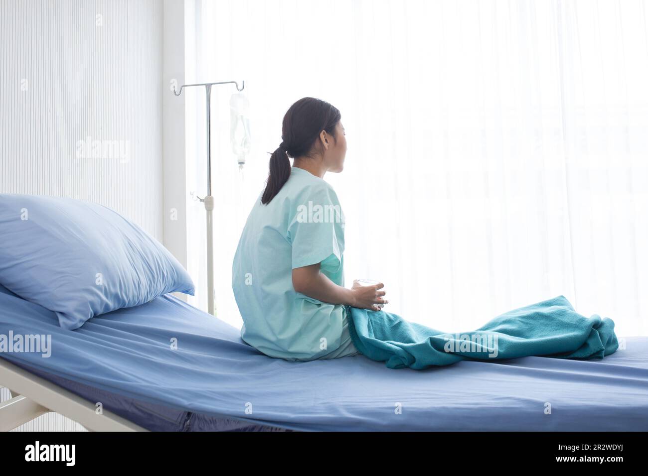 Eine asiatische Patientin saß auf einem Krankenhausbett mit ihrer rechten Hand und hielt ein Glas Wasser und es gibt eine Salzwasserlinie auf der linken Seite. Stockfoto