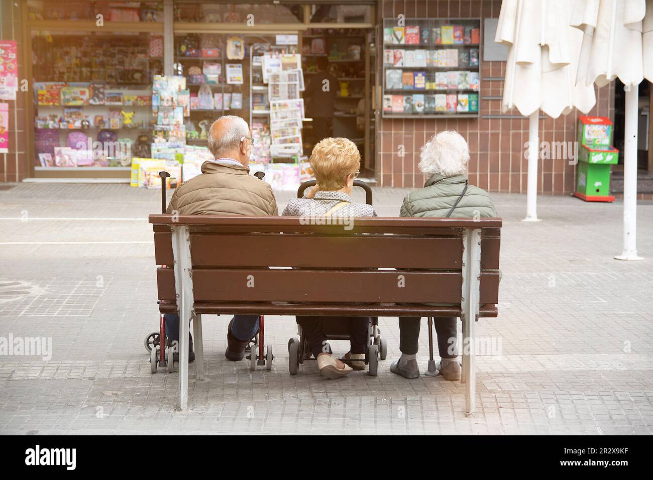 Drei ältere Menschen mit Mobilitätshilfen, die auf einer öffentlichen Straßenbank sitzen. Leerer Kopierbereich für Editortext. Stockfoto