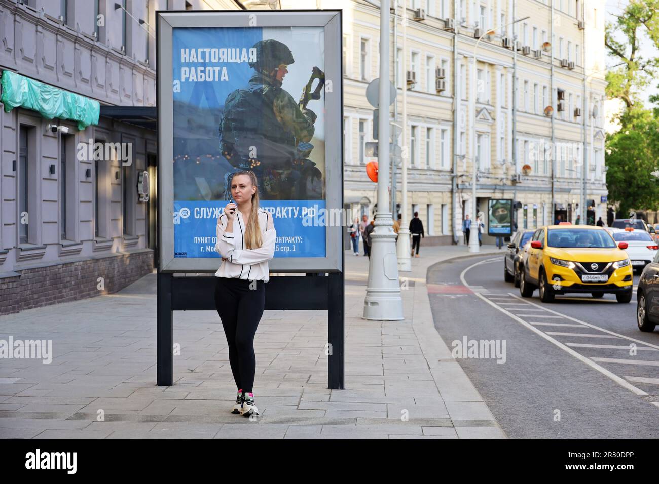 Ein Mädchen, das neben dem Plakat steht, mit einem Aufruf für Vertragsdienst bei der russischen Armee auf der Straße der Stadt Stockfoto