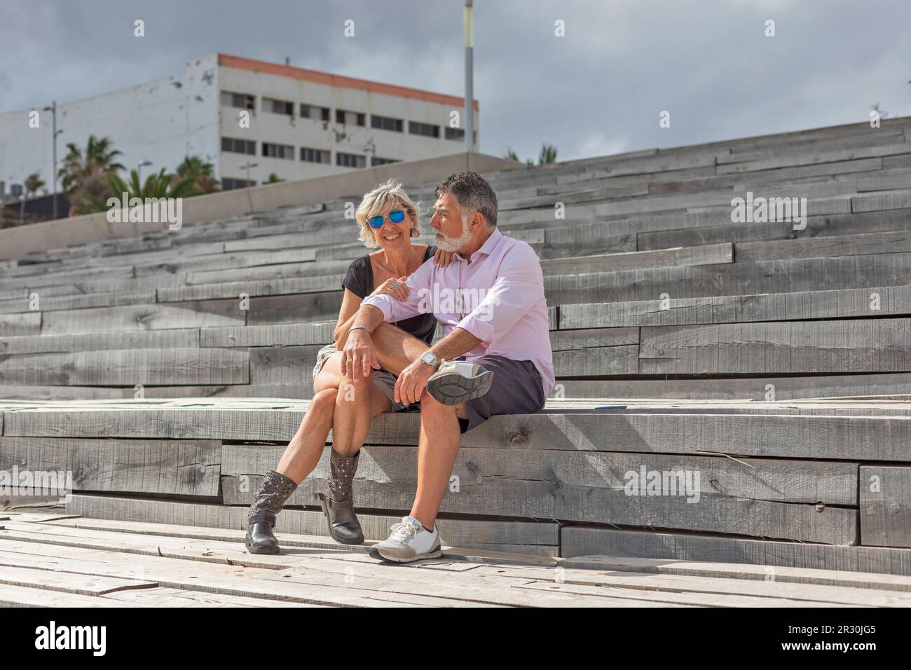 Ein glückliches Paar mittleren Alters lächelt, wenn es auf einer Treppe sitzt. Er ist vergrillt, hat einen weißen Bart und trägt eine Sonnenbrille. Sie lächelt und schaut in die Stockfoto