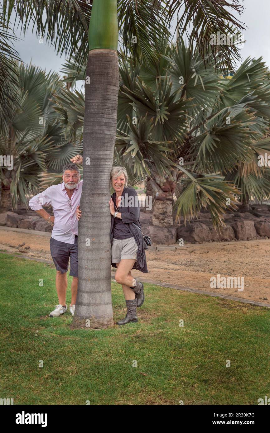 Ein heterosexuelles Paar in den 50ern macht Witze und lacht, wenn es sich an den Stamm einer Palme lehnt. Er verdreht seine Augen im Scherz. Sie trägt graue Shorts Stockfoto