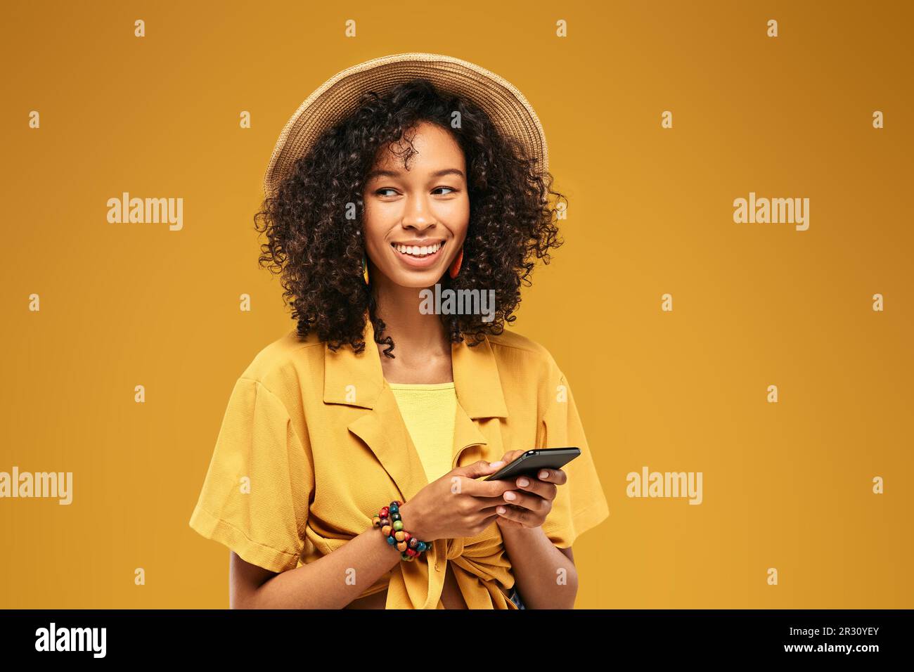Eine afroamerikanische Frau, die auf ihrem Smartphone eine Nachricht schreibt und lustige, gekleidete Strohmütze und Jugendkleidung auf gelbem Hintergrund trägt Stockfoto