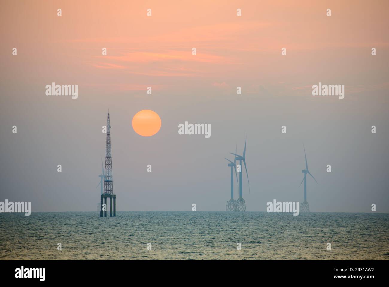 Fans von Windturbinen drehen sich über dem glitzernden Meer. Die orange-rote Sonne in der Dämmerung. Eine Offshore-Windfarm vor der Nordwestküste Taiwans. Einer von Green P. Stockfoto