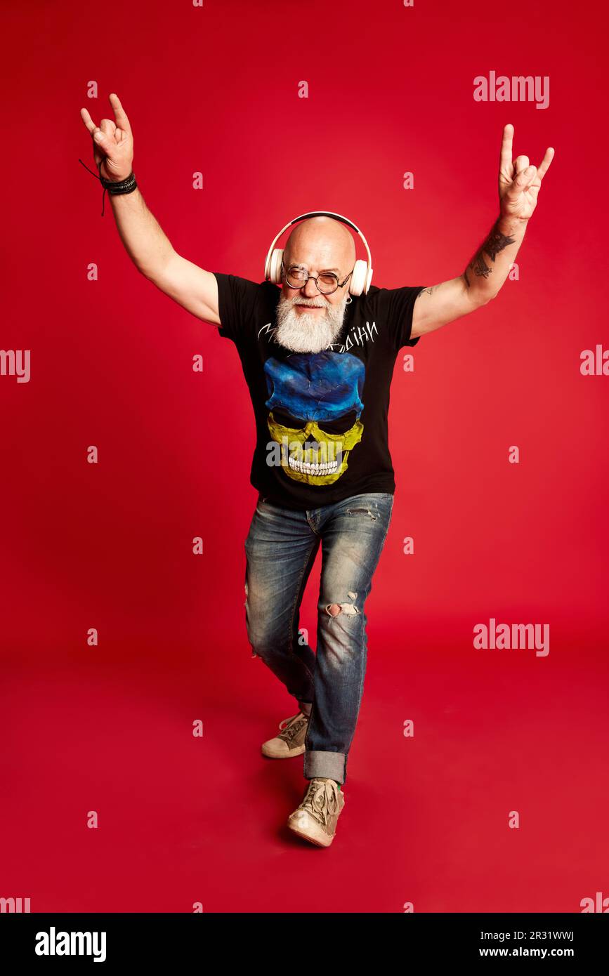 Rock 'n' Roll. Porträt in voller Länge emotionaler, reifer, bärtiger Mann mit Tattoos, der Musik in Kopfhörern vor rotem Studiohintergrund hört Stockfoto