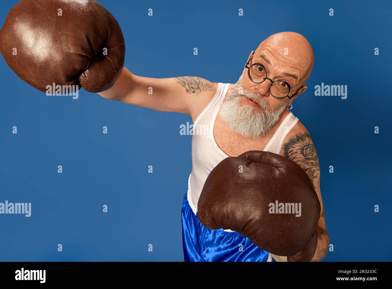 Porträt eines kahlköpfigen, bärtigen, reifen Mannes mit Tattoos in Sportbekleidung, der in alten Boxhandschuhen vor blauem Studiohintergrund posiert Stockfoto