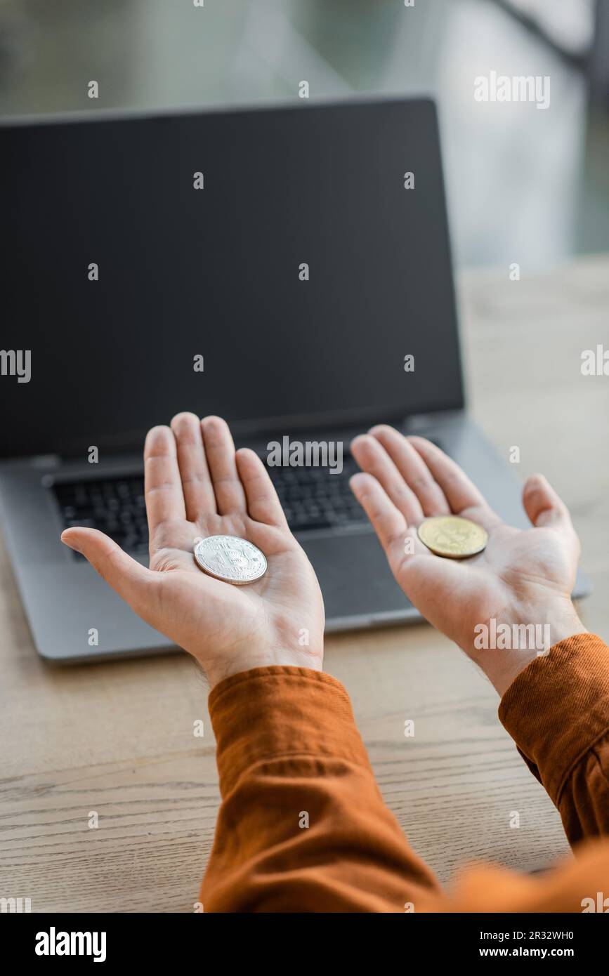 KIEW, UKRAINE - 18. OKTOBER 2022: Zugeschnittener Blick auf einen erfolgreichen Unternehmer, der goldene und silberne Bitcoins auf offenen Palmen in der Nähe eines Laptops mit einem leeren sc hält Stockfoto