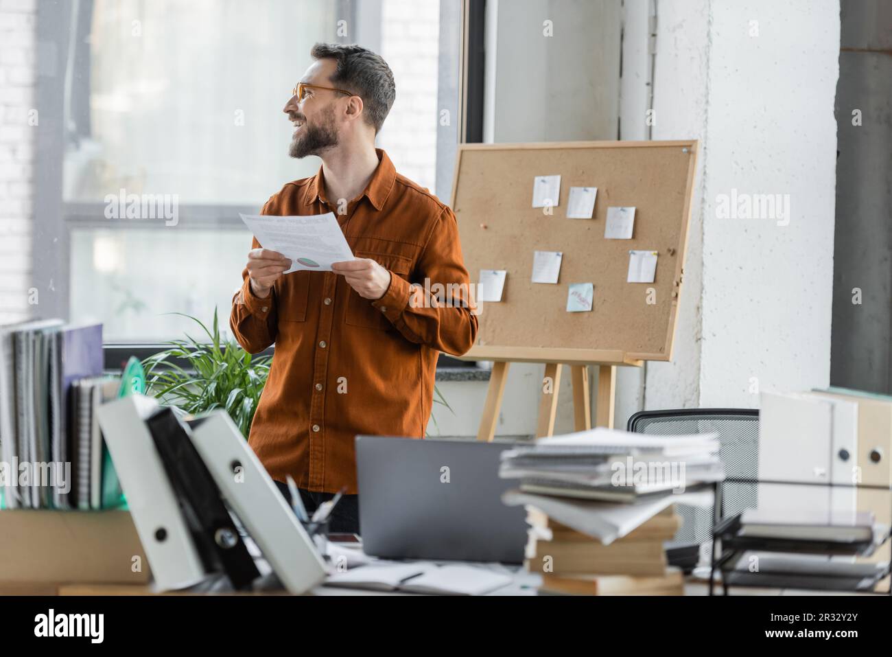 Ein positiver, erfolgreicher Unternehmer, der ein Dokument in der Hand hält und in die Nähe von Korkplatten mit Haftnotizen und Arbeitstisch mit Laptop, Ordnern, Büchern und nicht blickt Stockfoto