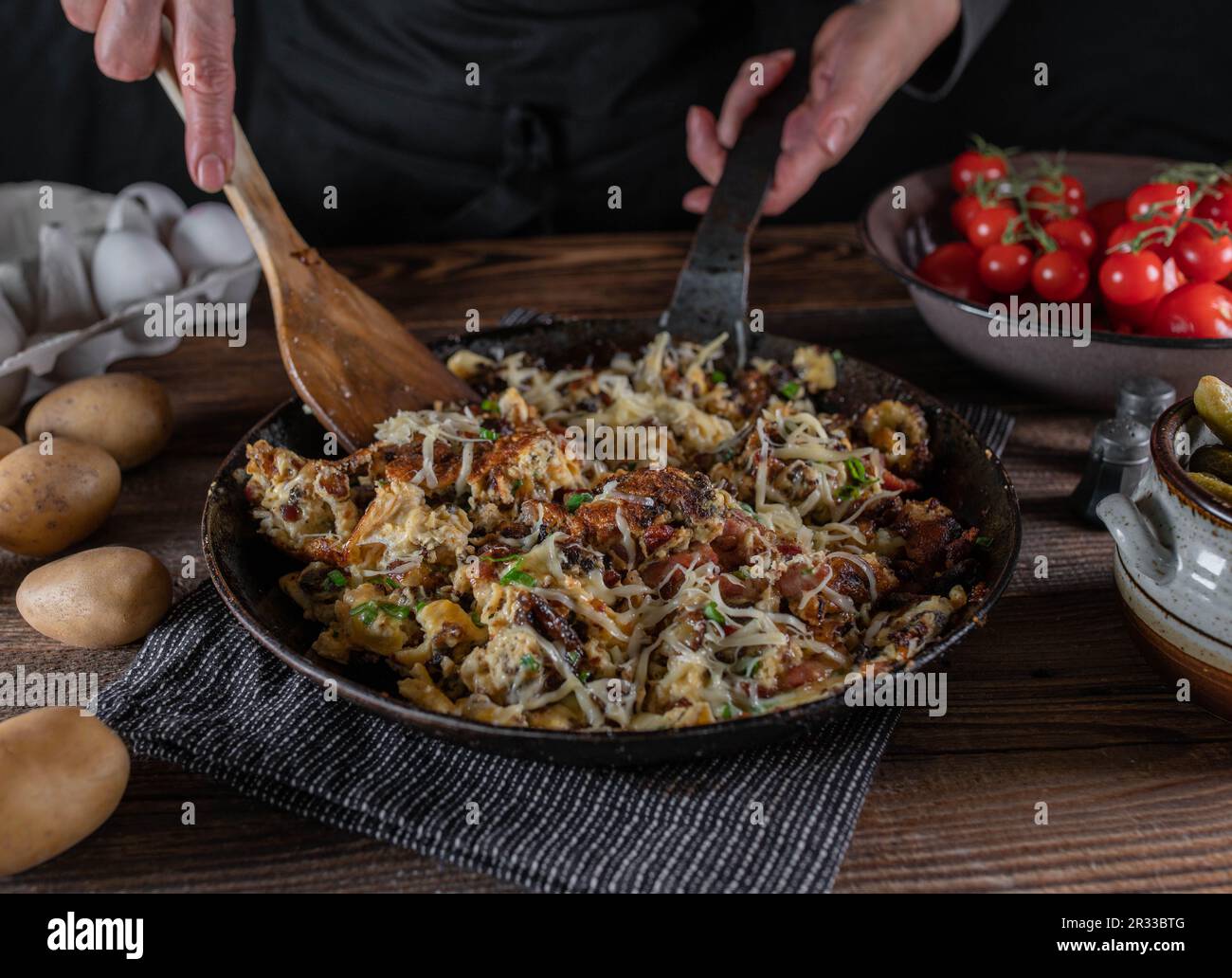 Bauernfrühstück mit Bratkartoffeln, Speck, Eiern, Schnittlauch und Käse. Serviert in einer rustikalen Eisenpfanne auf einem Holztisch. Stockfoto