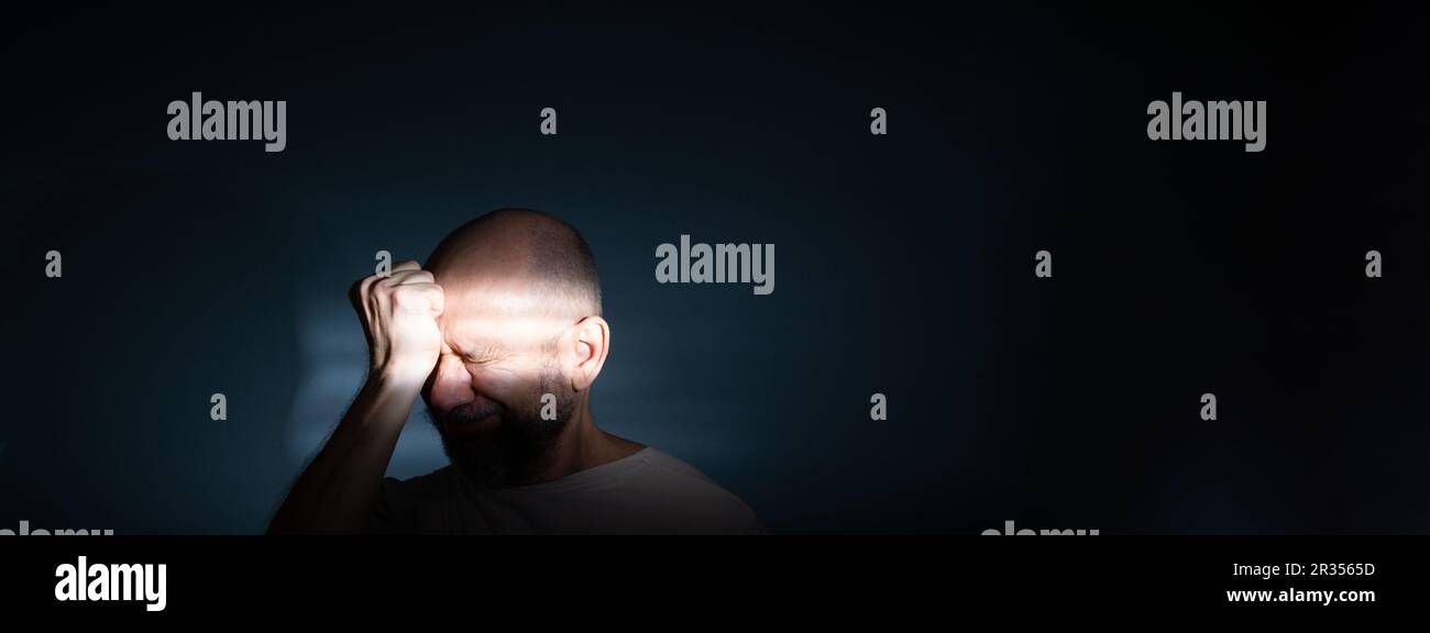 Trauriger Mann mit Kopfschmerzen oder Migräne. Gestresster Kerl mit schmerzhaftem Gesichtsausdruck, der sich schrecklich schwach oder depressiv fühlt. Isoliert auf dunklem Bac Stockfoto