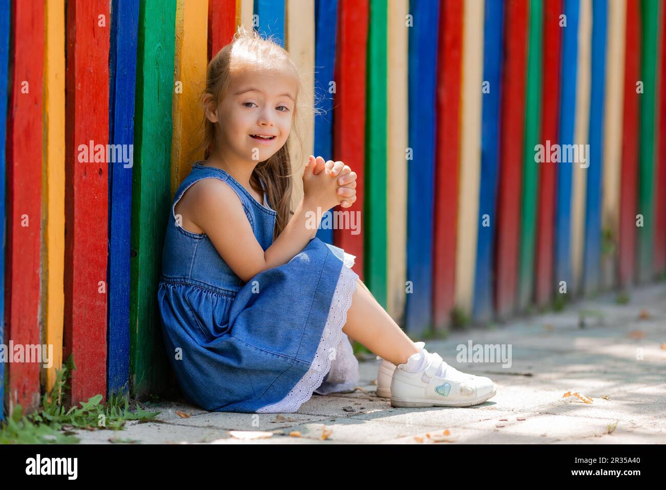 Ein kleines Mädchen mit Daunensyndrom, das eine Brille zum Sehen trägt, sitzt im Sommer neben einem farbigen Zaun in einer Kinderstadt. Hochwertiges Foto Stockfoto