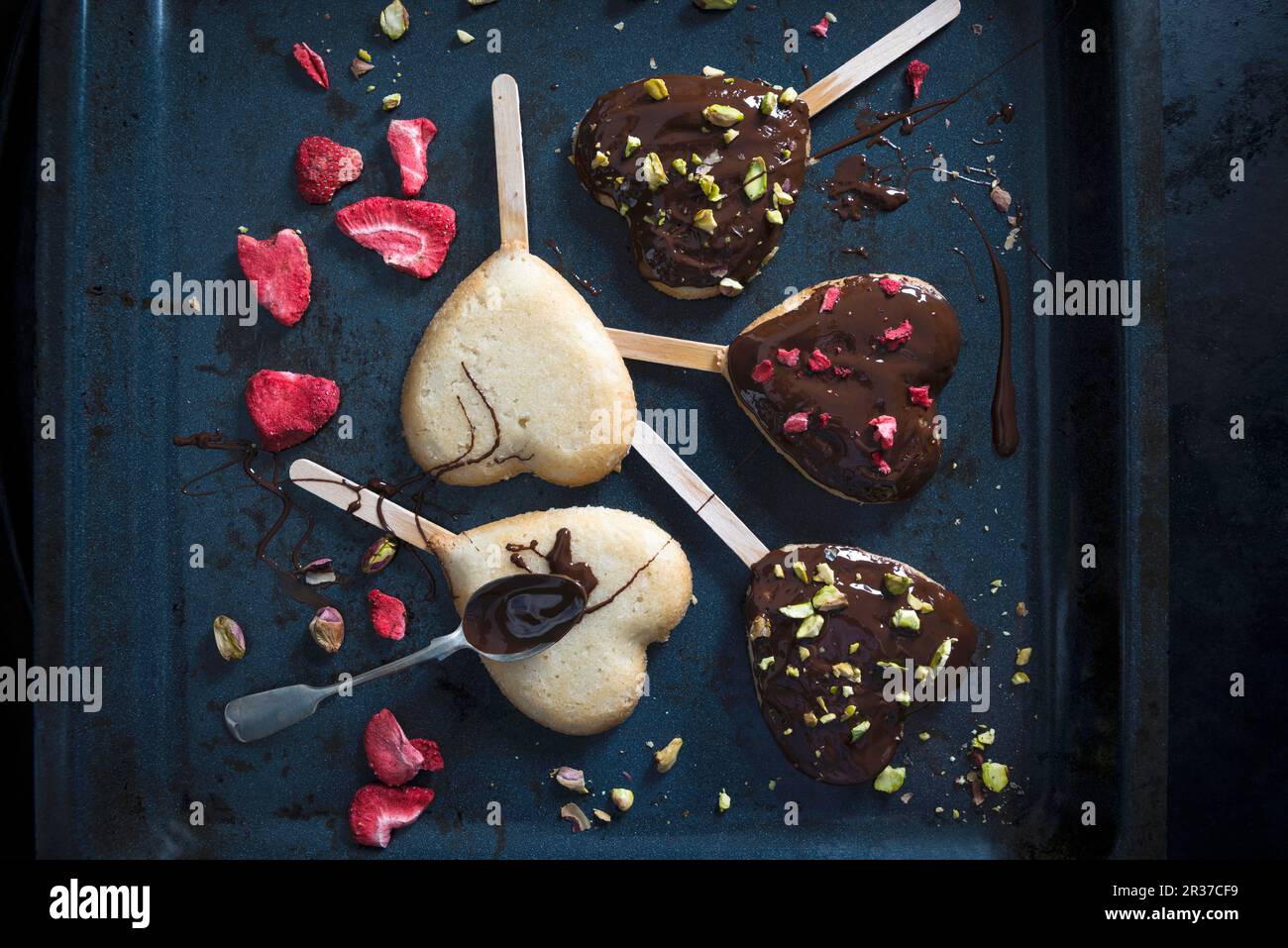 Herzförmige vegane Kuchen auf Stöcken, dekoriert mit einer dunklen Bierglasur, gefriergetrockneten Erdbeeren und Pistazien Stockfoto