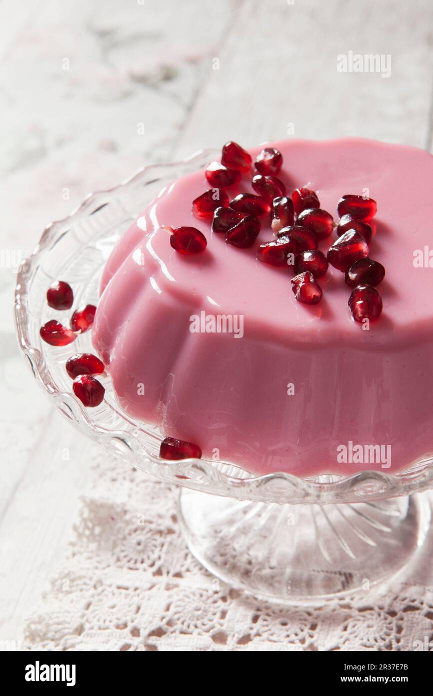Rosafarbenes Milchgelee mit Granatapfelsamen auf einem Kuchenständer aus Glas und klassischer Tischdecke aus Spitze Stockfoto