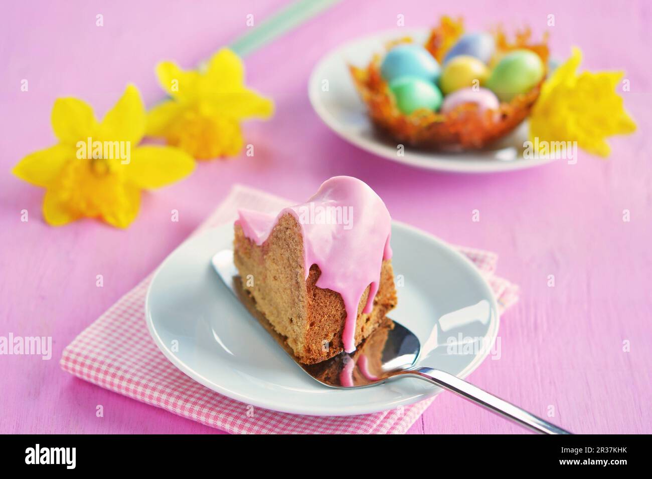 Ein Stück Gugelhupf mit rosafarbener Zuckerglasur, ein Karamellnest mit bunten Zuckereiern und Narzissen im Hintergrund Stockfoto