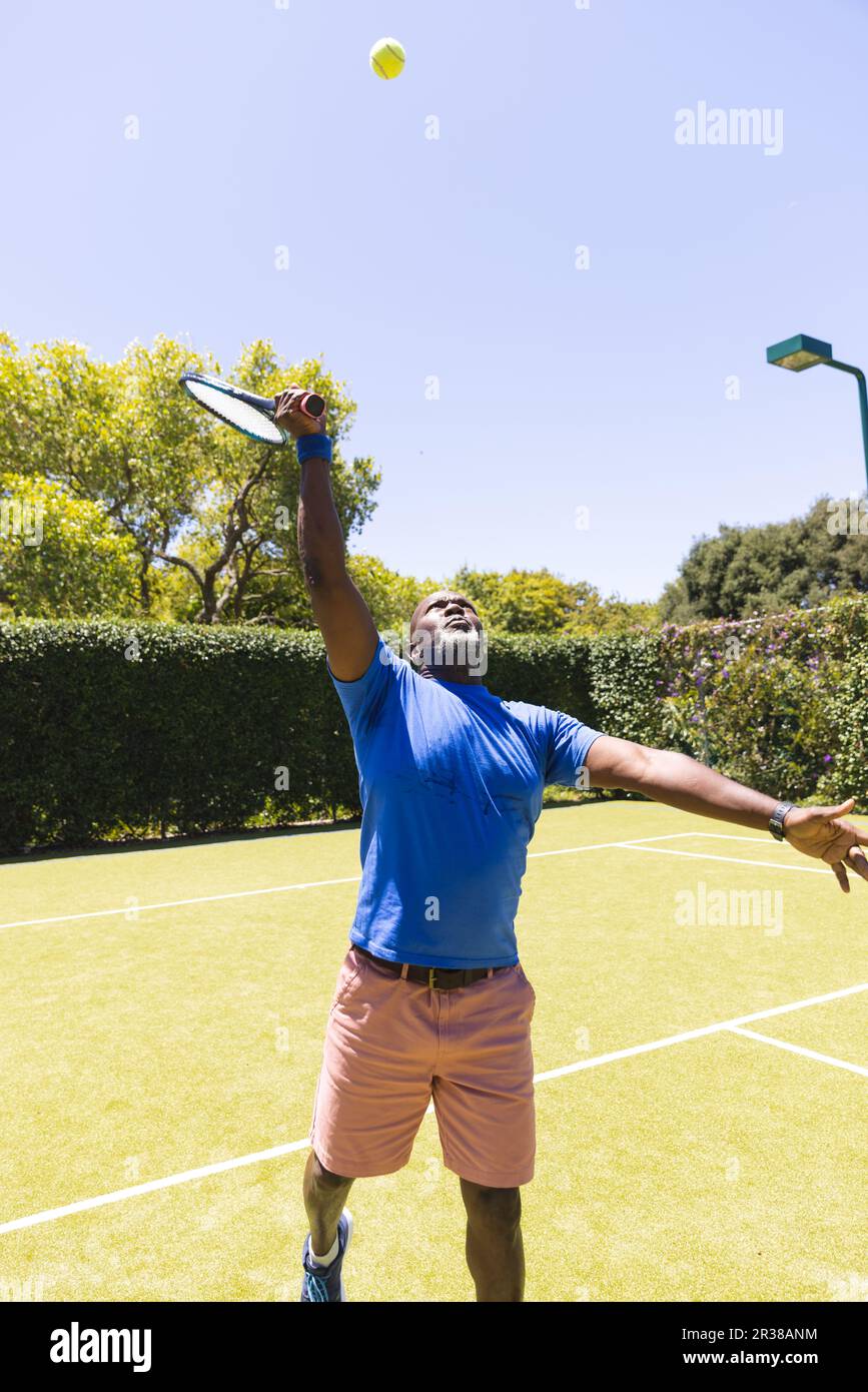 Glücklicher afroamerikanischer Senior-Mann, der Tennis spielt und sich streckt, um High Ball auf einem sonnigen Rasenplatz zu schlagen Stockfoto