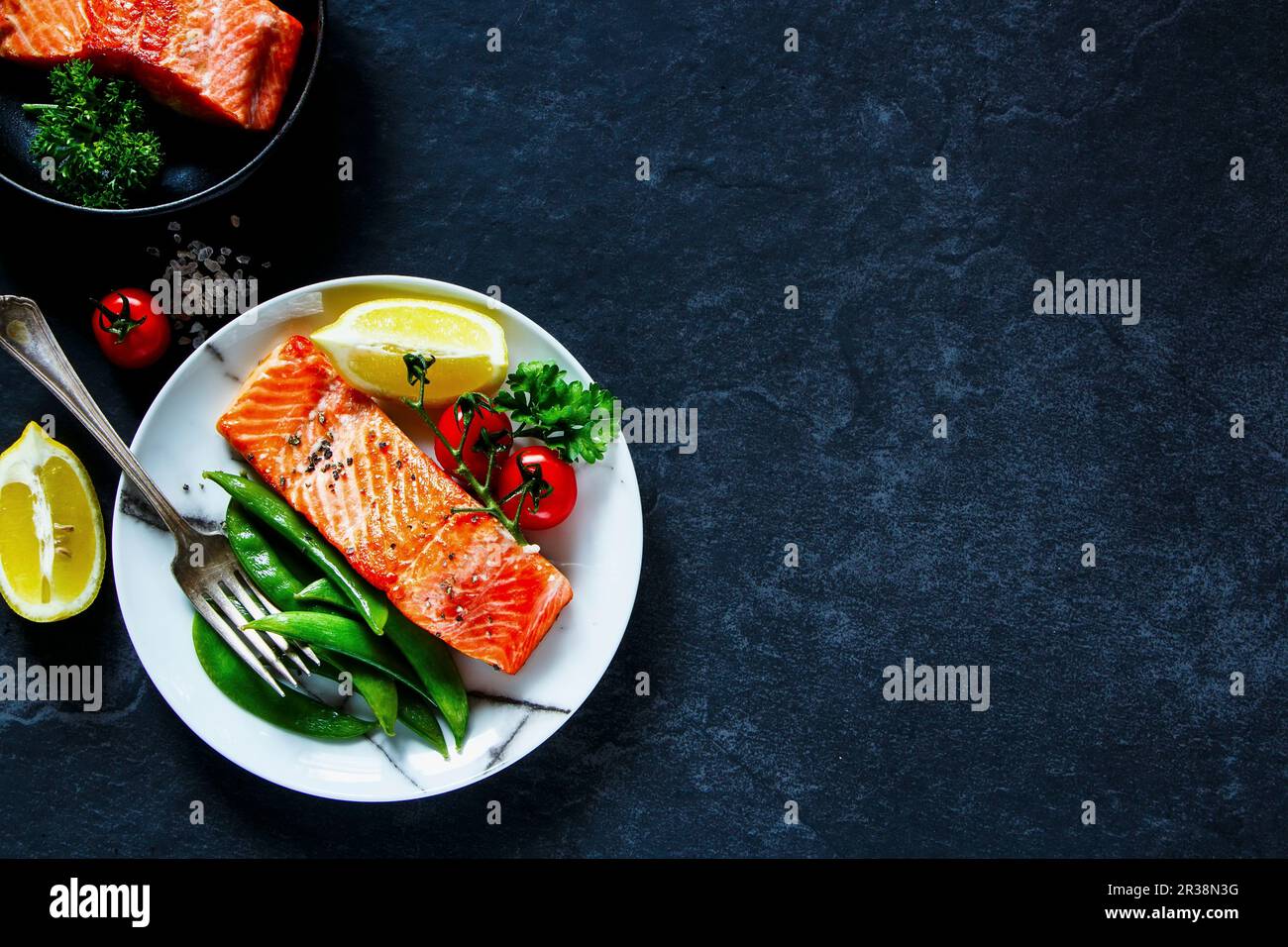Gerösteter Lachs mit pochierten grünen Erbsen, Tomaten, Zitrone auf dem Teller und Lachsfilet in klassischer Bratpfanne Stockfoto