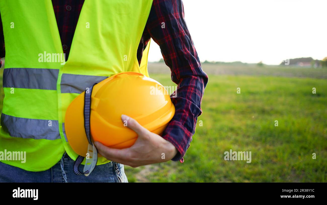 Bauarbeiter mit seinem gelben Helm, Schutzhelm und Bauplan trägt fluoreszierende Weste auf dem leeren Feld. OSH. Stockfoto