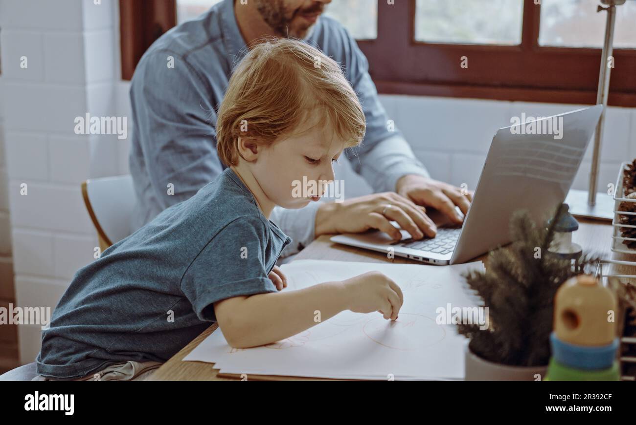 Vater, der im Home Office mit einem Laptop arbeitet, sitzt am Tisch, und der süße kleine Sohn spielt, um ihn zu stören. Stockfoto