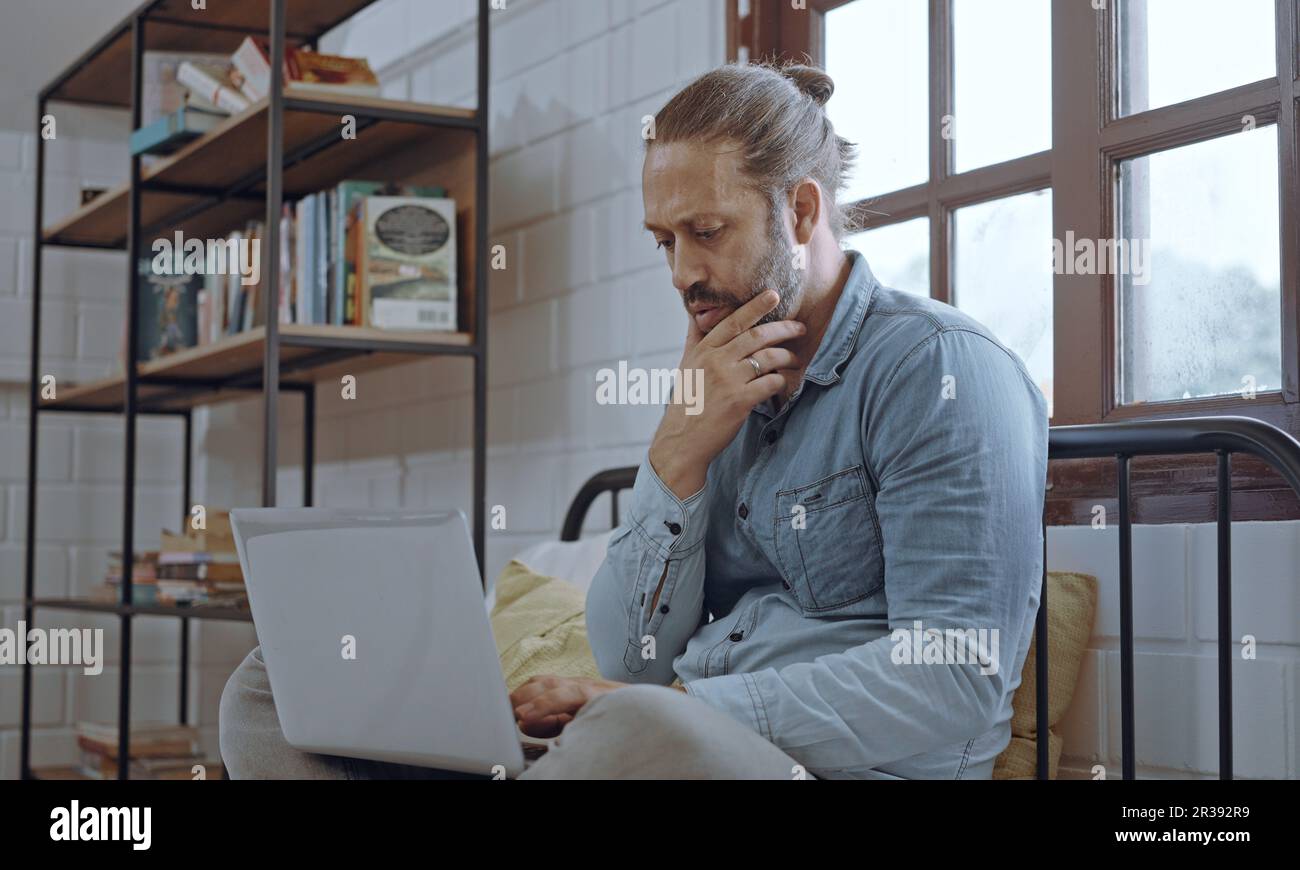 Ein junger besorgter Mann, der ein Notebook benutzt und im Schlafzimmer auf dem Bett sitzt. Stockfoto