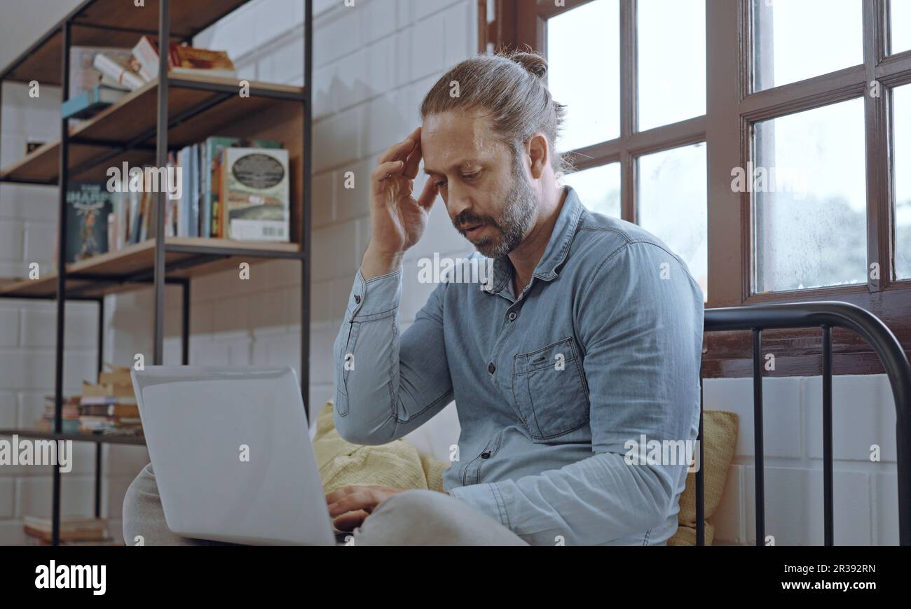 Ein junger besorgter Mann, der ein Notebook benutzt und im Schlafzimmer auf dem Bett sitzt. Stockfoto