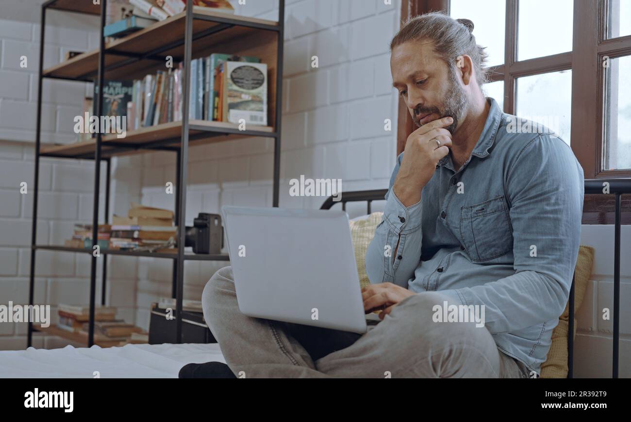 Ein junger besorgter Mann, der ein Notebook benutzt und im Schlafzimmer auf dem Bett sitzt. Stockfoto
