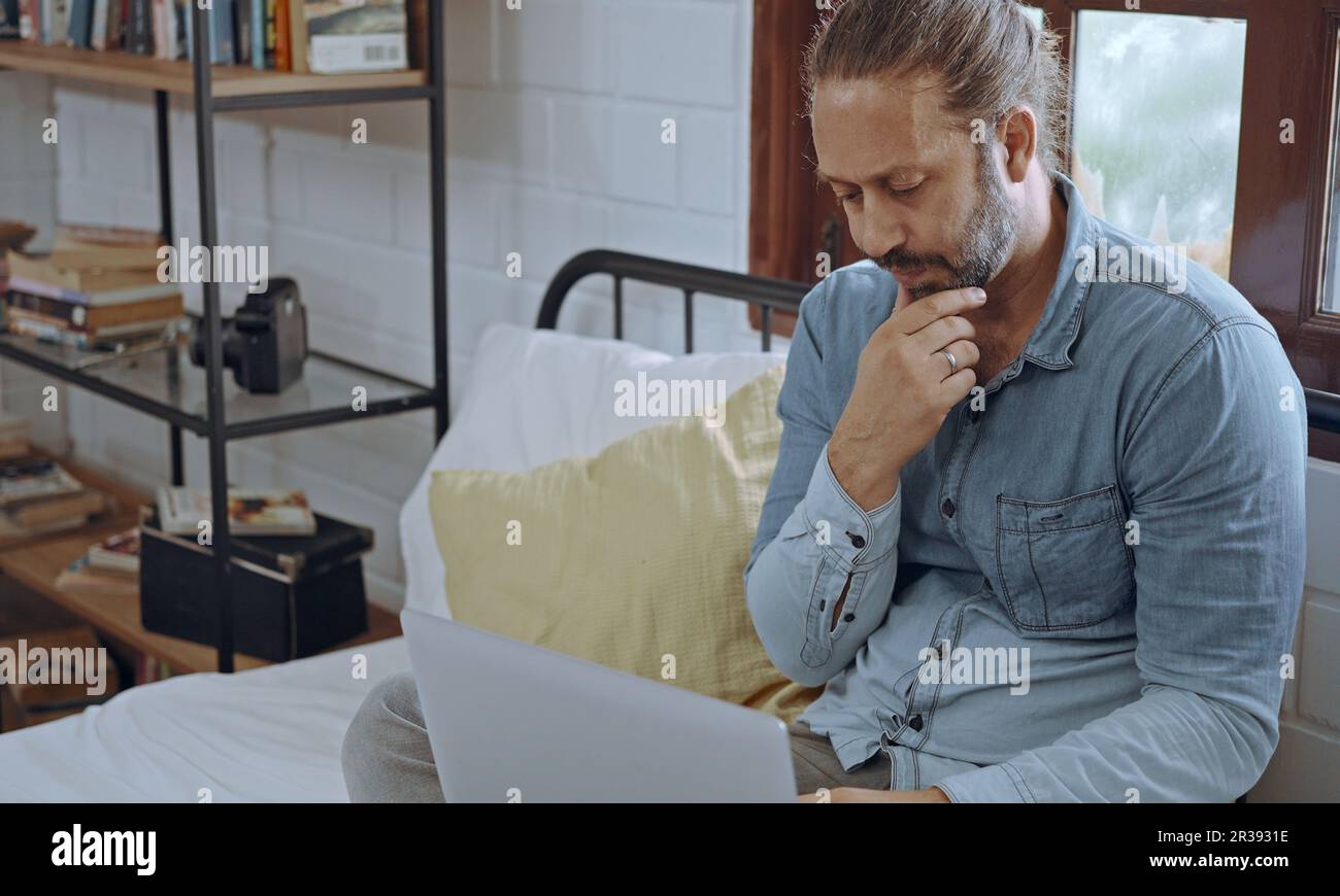 Ein junger besorgter Mann, der ein Notebook benutzt und im Schlafzimmer auf dem Bett sitzt. Stockfoto