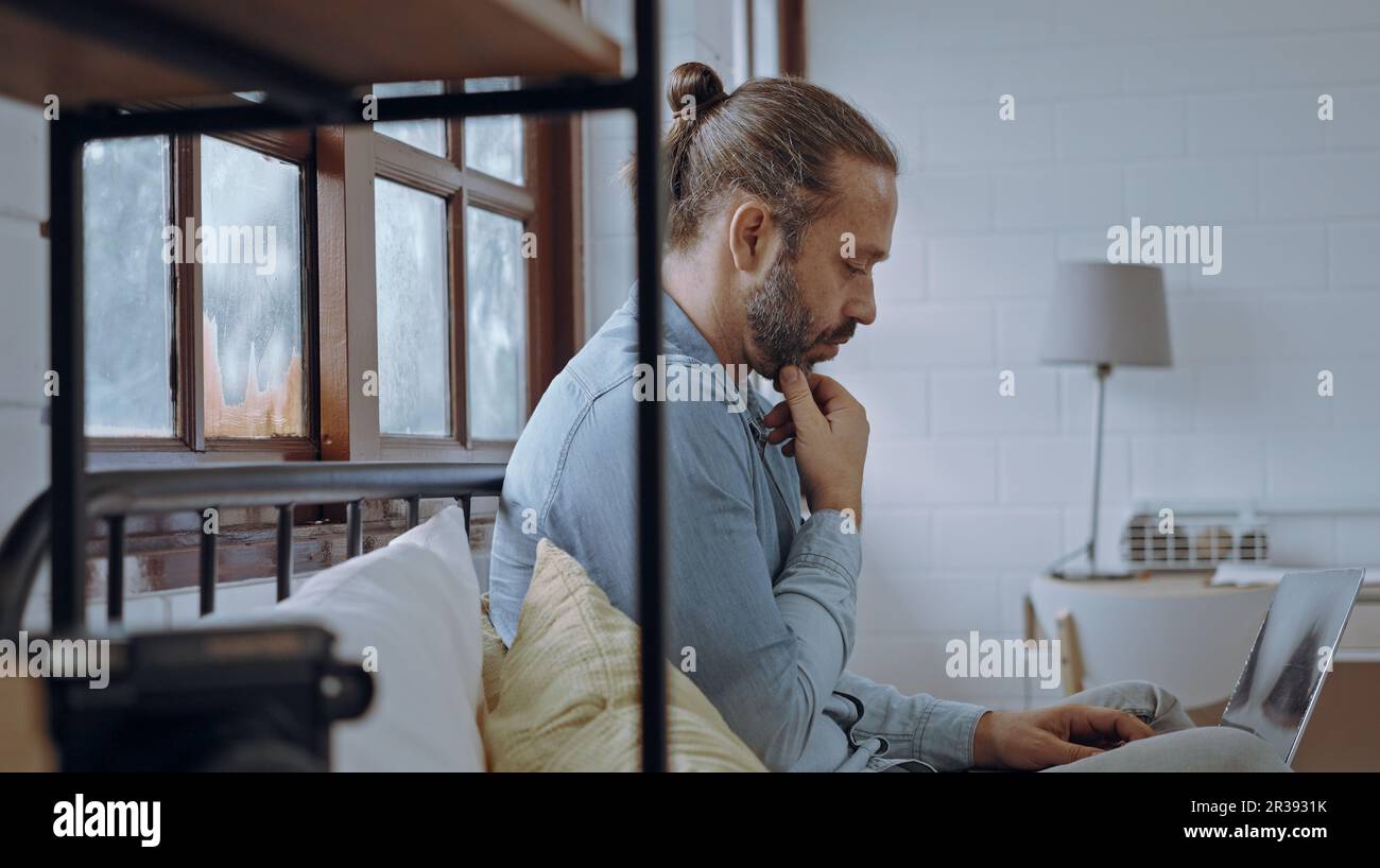 Ein junger besorgter Mann, der ein Notebook benutzt und im Schlafzimmer auf dem Bett sitzt. Stockfoto