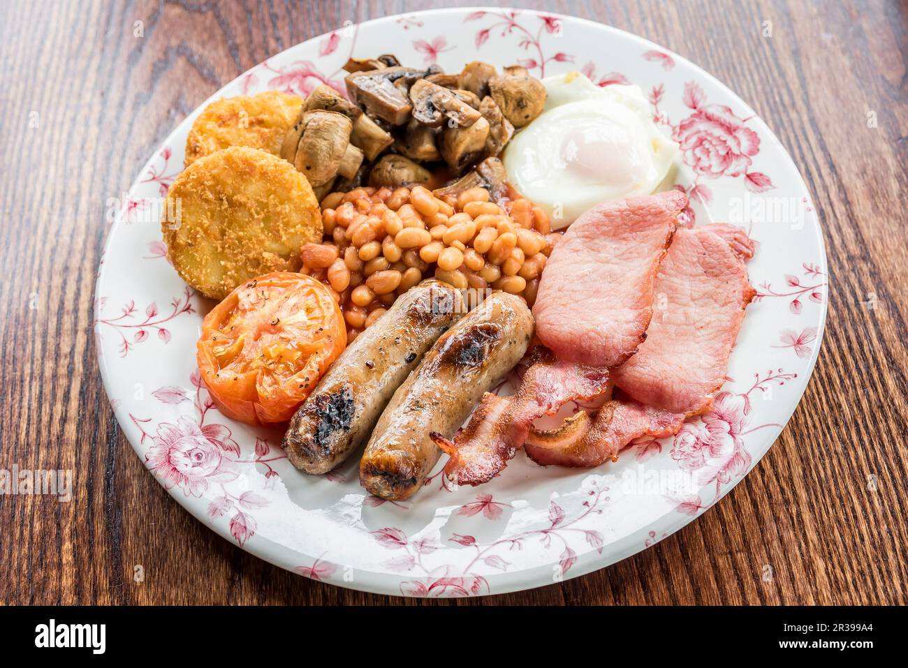 Traditionelles englisches Frühstück mit Speck, Würstchen, gegrillten Tomaten und Pilzen, gebackenen Bohnen, Kartoffelrösti Kuchen und Ei auf einem Holztisch Stockfoto