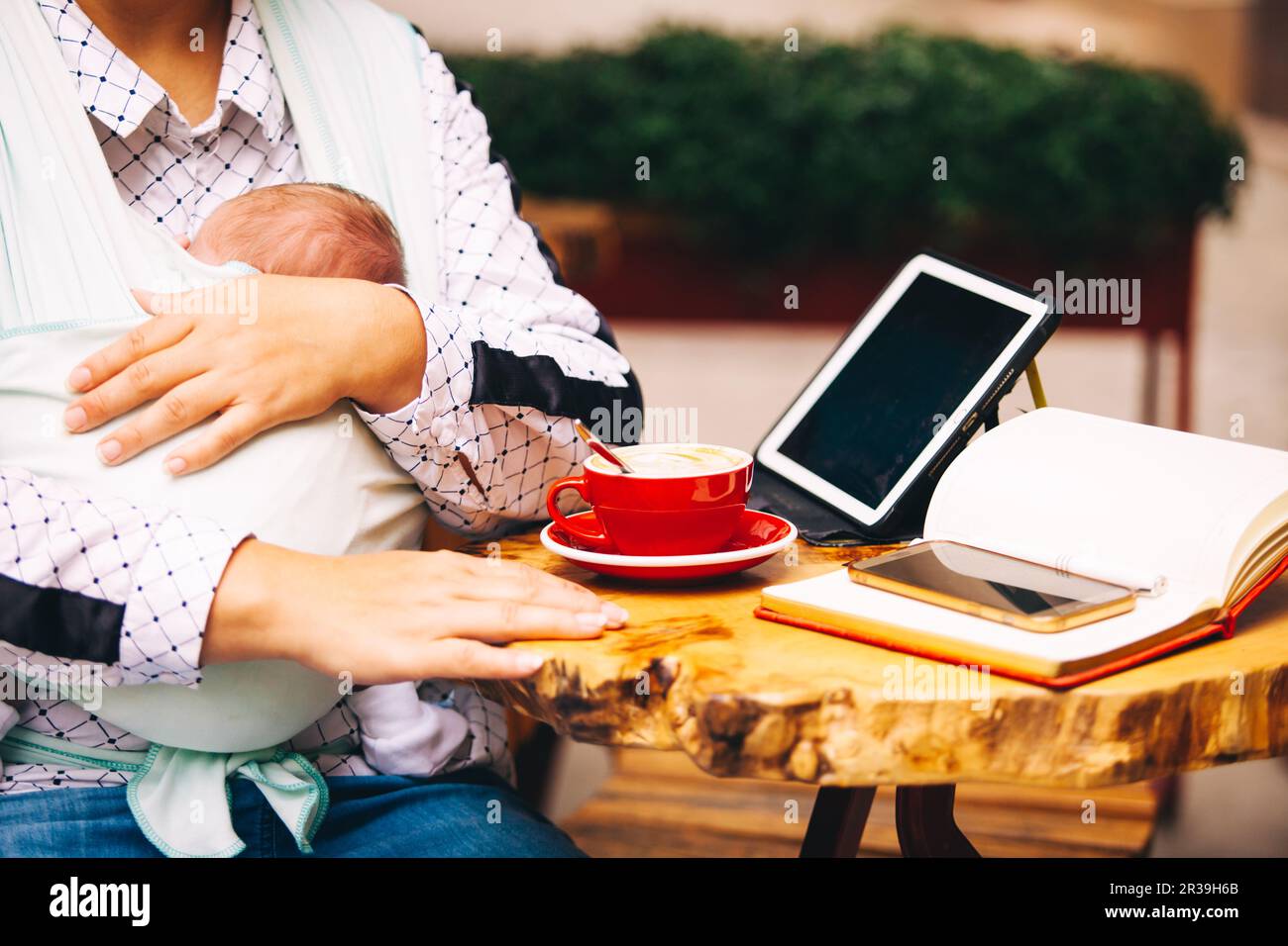 Freiberufler, der mit dem Neugeborenen im Freien arbeitet. Frau, die ein Tablet benutzt, wenn das Baby in der Schlinge schläft Stockfoto